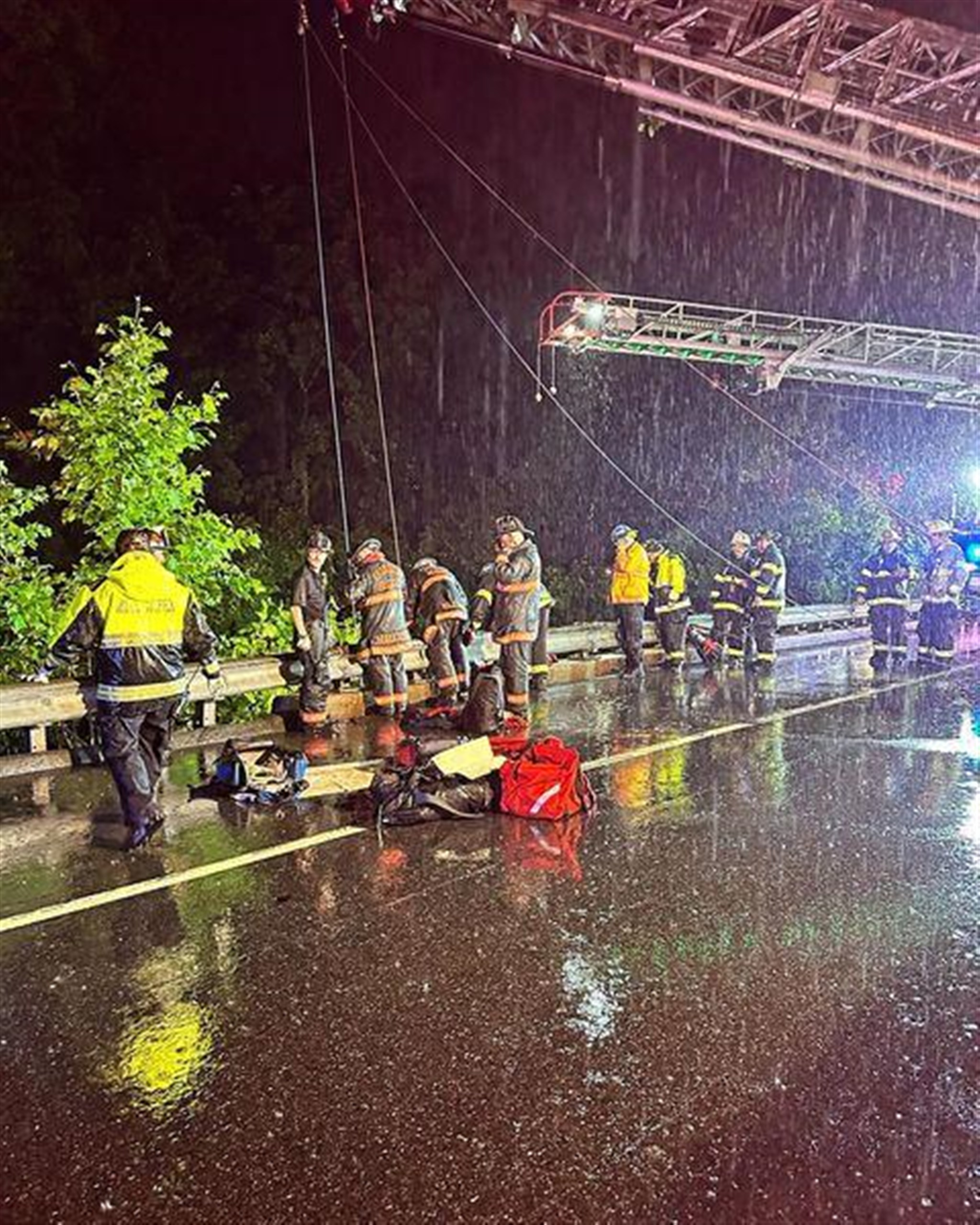 A group of firefighters and rescue workers respond to an emergency on a rain-soaked road at night. They are gathered near a damaged guardrail, with emergency lights illuminating the scene. Equipment and gear are scattered on the wet pavement.
