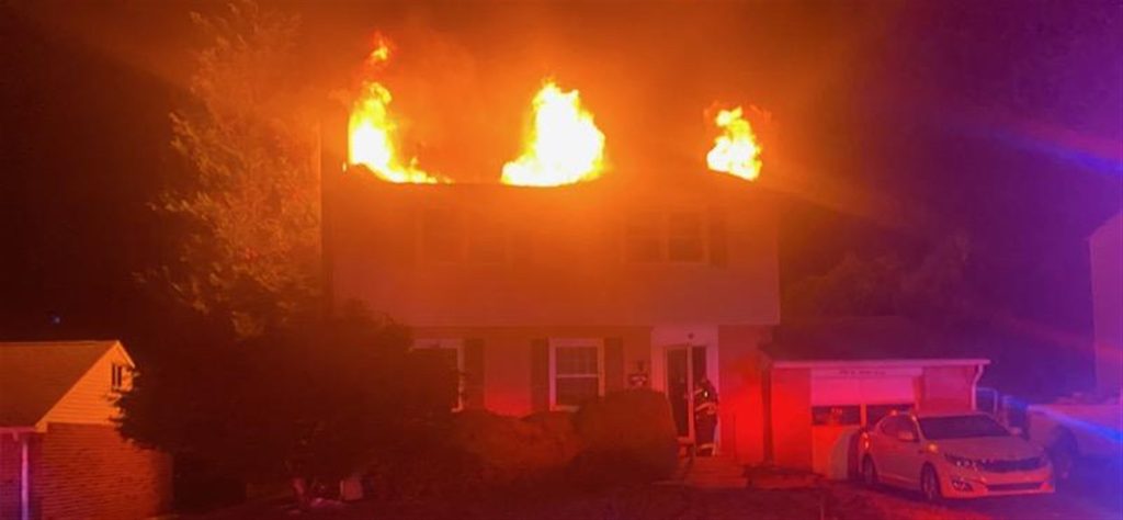 A two-story house is engulfed in flames on the upper floor during nighttime. Bright orange fire illuminates the roof area, and smoke is visible. A car is parked in the driveway, and the scene is lit by emergency response lights.