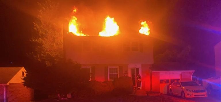A two-story house is engulfed in flames on the upper floor during nighttime. Bright orange fire illuminates the roof area, and smoke is visible. A car is parked in the driveway, and the scene is lit by emergency response lights.
