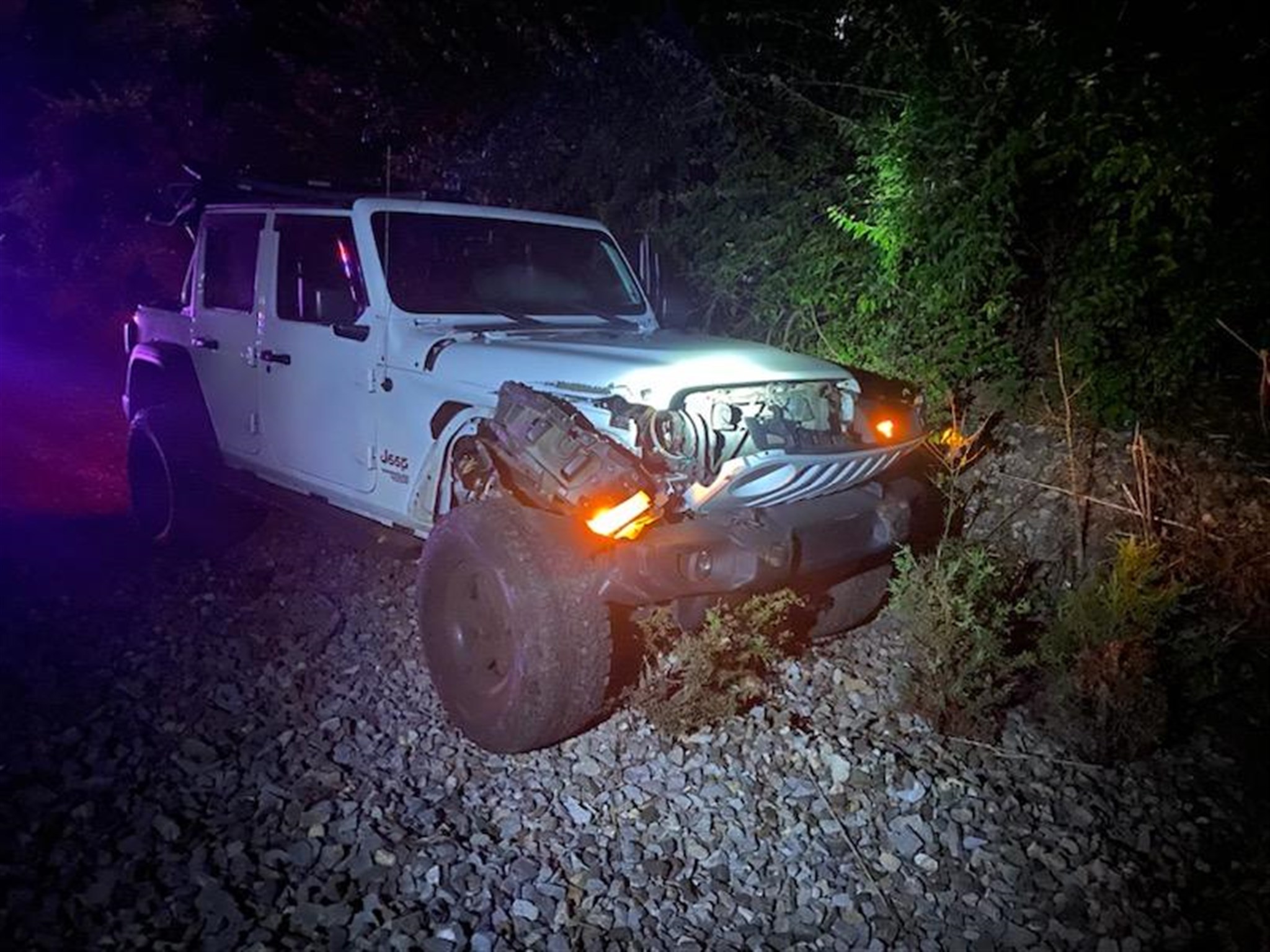 A white Jeep with significant front-end damage is situated off-road in a forested area at night. The headlights are on, illuminating the surrounding foliage, and parts of the vehicle's front are visibly distorted, suggesting a recent collision.