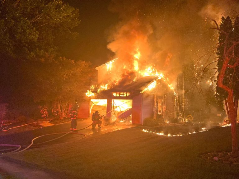 A house engulfed in flames during the night. Several firefighters are working to extinguish the fire with hoses. The scene is brightly lit by the blaze, and smoke is billowing into the sky. Trees are visible on either side of the burning house.