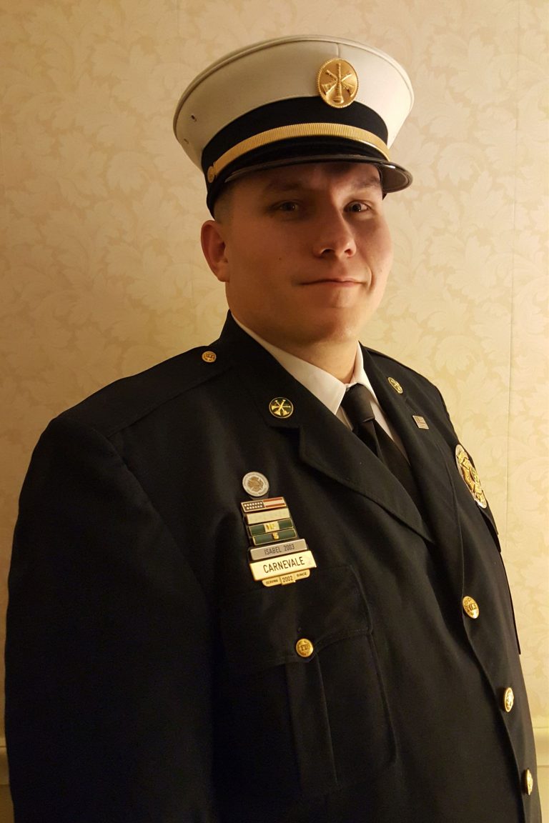 A person in a dark formal uniform with various pins and a name badge stands against a patterned beige wall. They are wearing a peaked cap with a badge at the center and looking towards the camera with a slight smile.