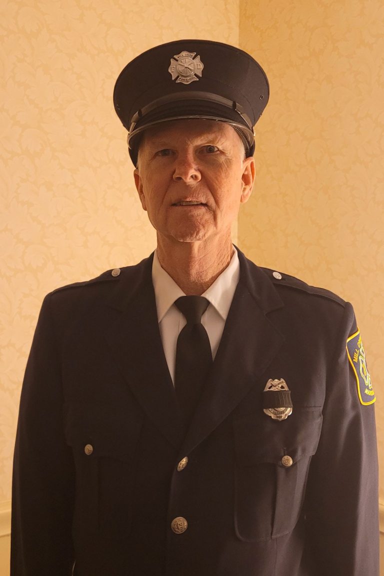 A person standing indoors wearing a dark blue fire department uniform with a matching hat. The uniform includes a badge on the chest and an insignia on the sleeve, as well as a white shirt and black tie underneath. The background features a light-colored, patterned wall.