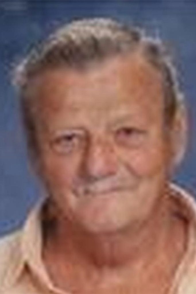 A portrait of an older man with light skin and gray hair. He is smiling slightly, wearing a beige shirt with the collar open. The background is a soft blue.