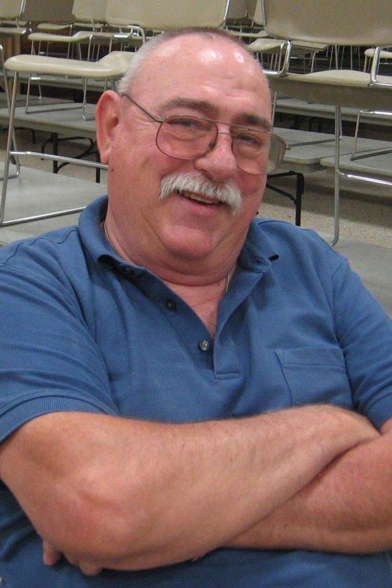 A man with a bald head, white mustache, and glasses smiles with his arms crossed. He is wearing a blue polo shirt and is seated in front of rows of empty chairs.