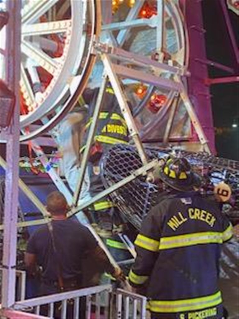 Firefighters work to rescue a person trapped in a Ferris wheel at night. The Ferris wheel is brightly lit, and several firefighters, wearing protective gear with "Mill Creek" on their uniforms, are assisting in the operation.
