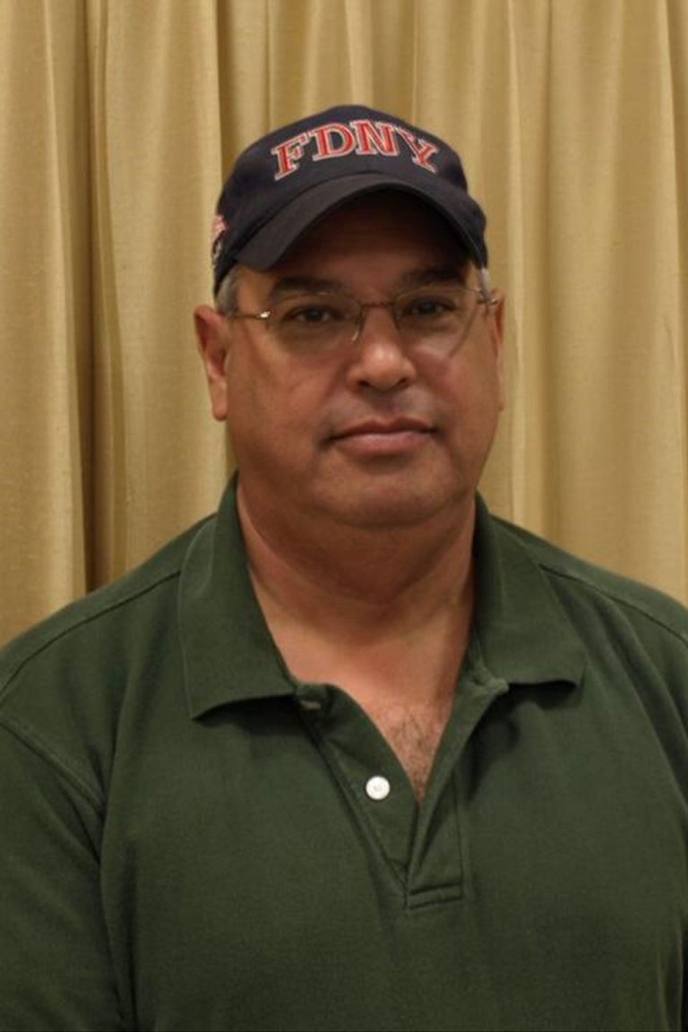 A person wearing a green polo shirt and an FDNY cap stands in front of a beige curtain. They have short hair, glasses, and a neutral expression.