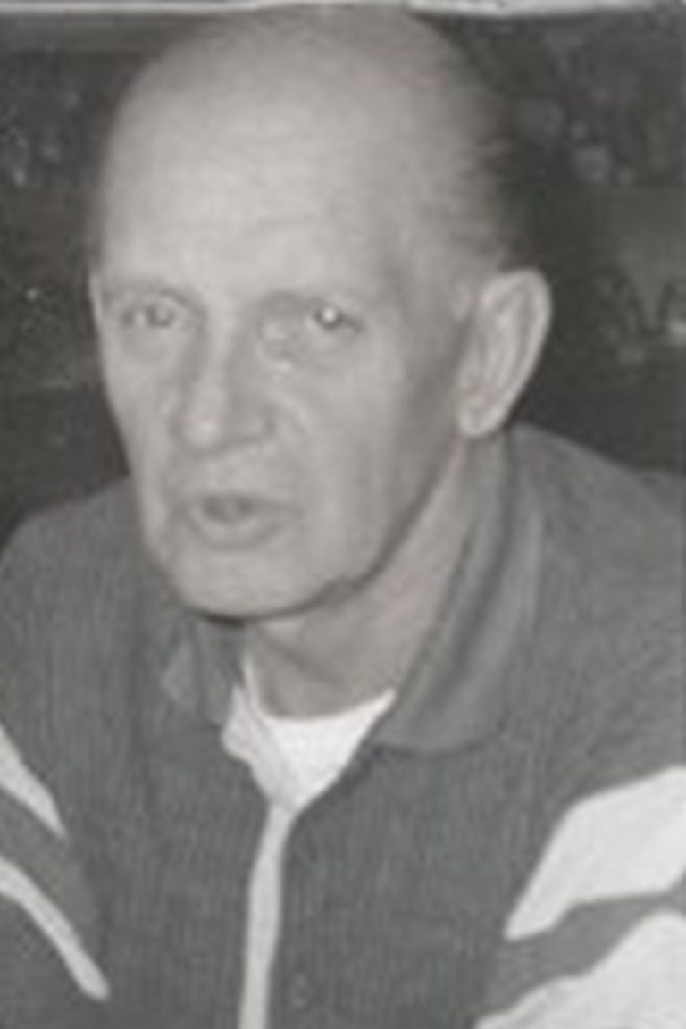 Black-and-white photo of an older man with a receding hairline. He is wearing a collared shirt with stripes on the sleeves and has a serious expression. The background is blurred, making it difficult to discern details.