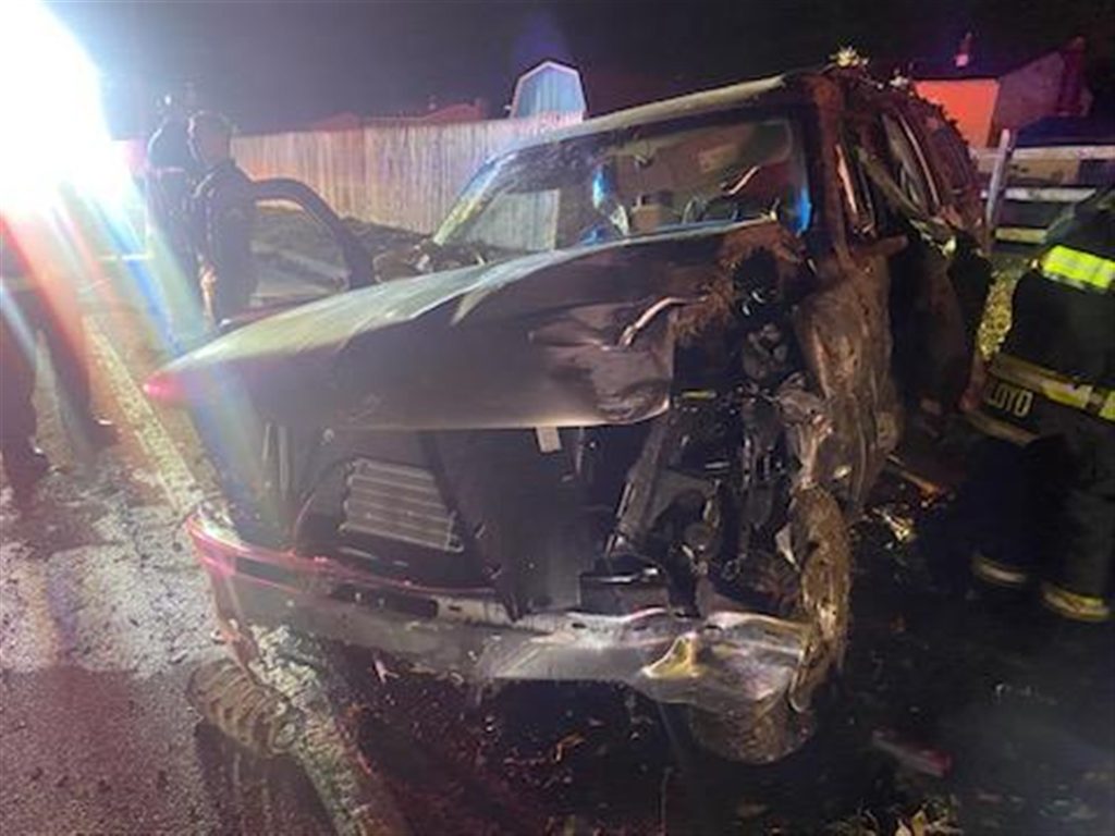 A severely damaged SUV is shown at night, with a crushed front and side. Emergency responders, including firefighters, stand near the vehicle, illuminated by bright lights from emergency vehicles in the background. The SUV appears to have collided with a fixed object.
