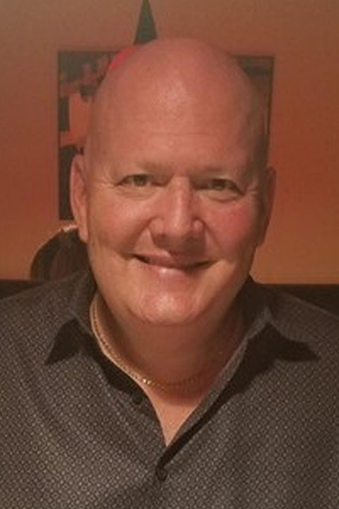 A smiling bald man with fair skin is wearing a dark button-up shirt with a subtle pattern. He is sitting indoors in a softly lit room, and a piece of abstract art is visible on the wall behind him.