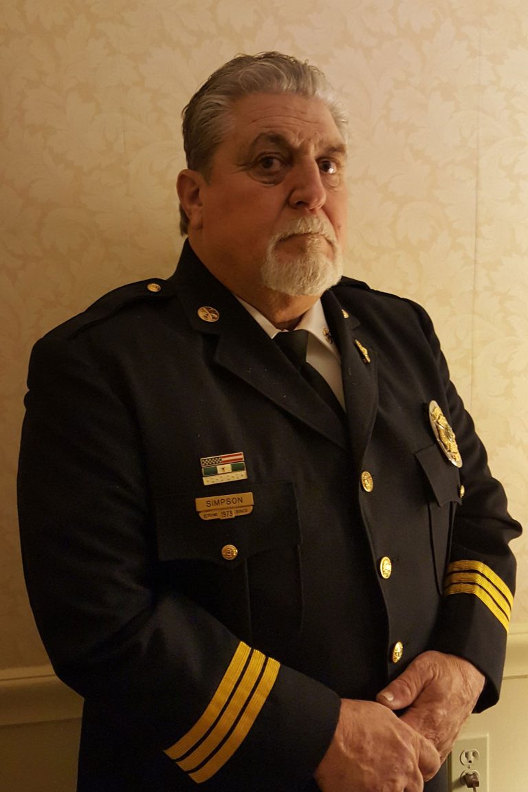 A mature man with a gray beard is standing in front of a light-colored patterned wall. He is wearing a dark formal uniform with gold stripes on the sleeves, a badge on the chest, and a name tag that reads "Simpson." His hands are clasped in front of him.
