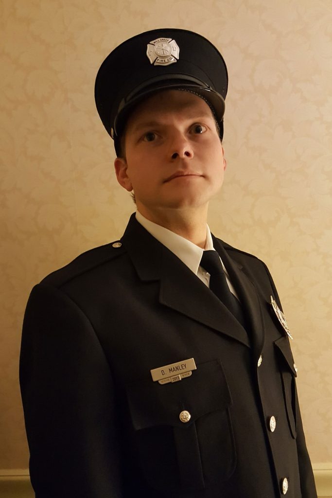 A person wearing a firefighter uniform and hat stands against a beige wallpapered background. They have a badge on the hat and a name tag on their chest that reads "D. Manley". They are looking directly into the camera with a serious expression.