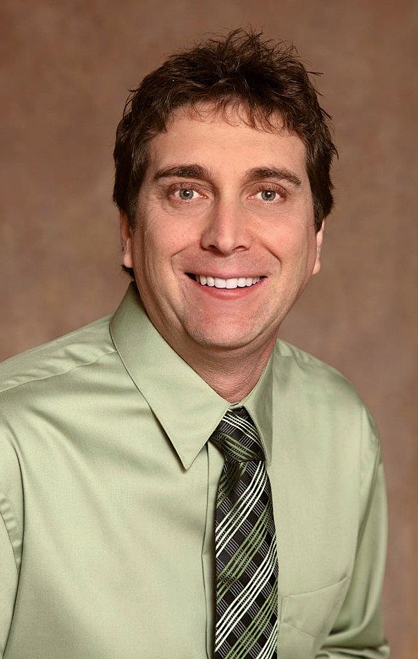 A man with short brown hair smiles at the camera. He is wearing a light green dress shirt and a patterned green tie. The background is a soft brown gradient.