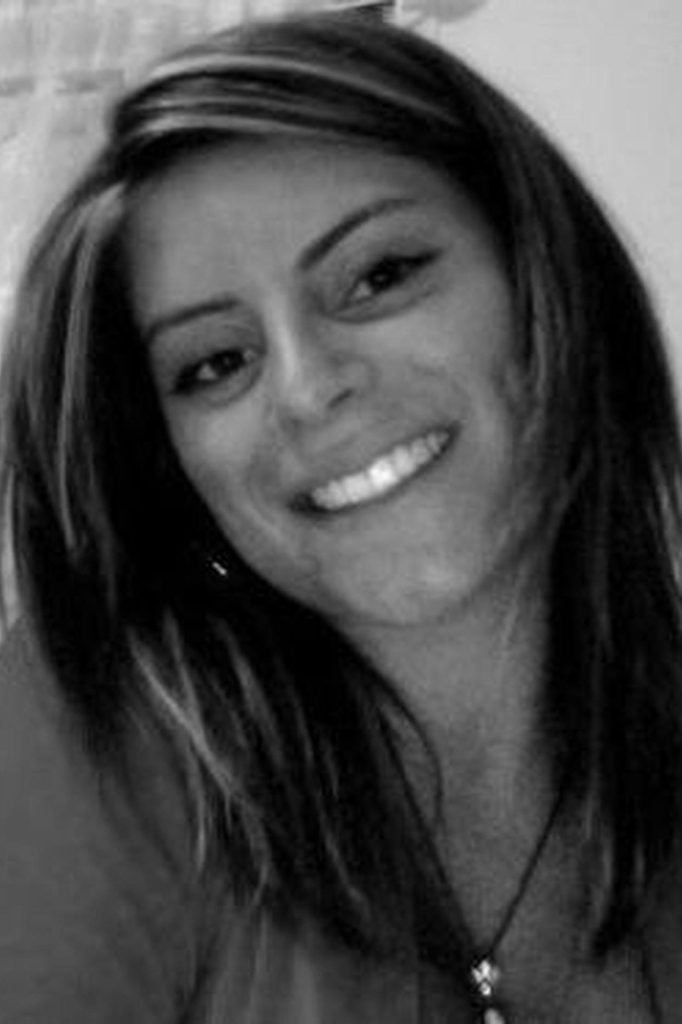 A grayscale photo of a woman with straight hair, smiling warmly at the camera. She has layered hair that falls past her shoulders and is wearing a necklace. The background is blurred, focusing attention on her face.