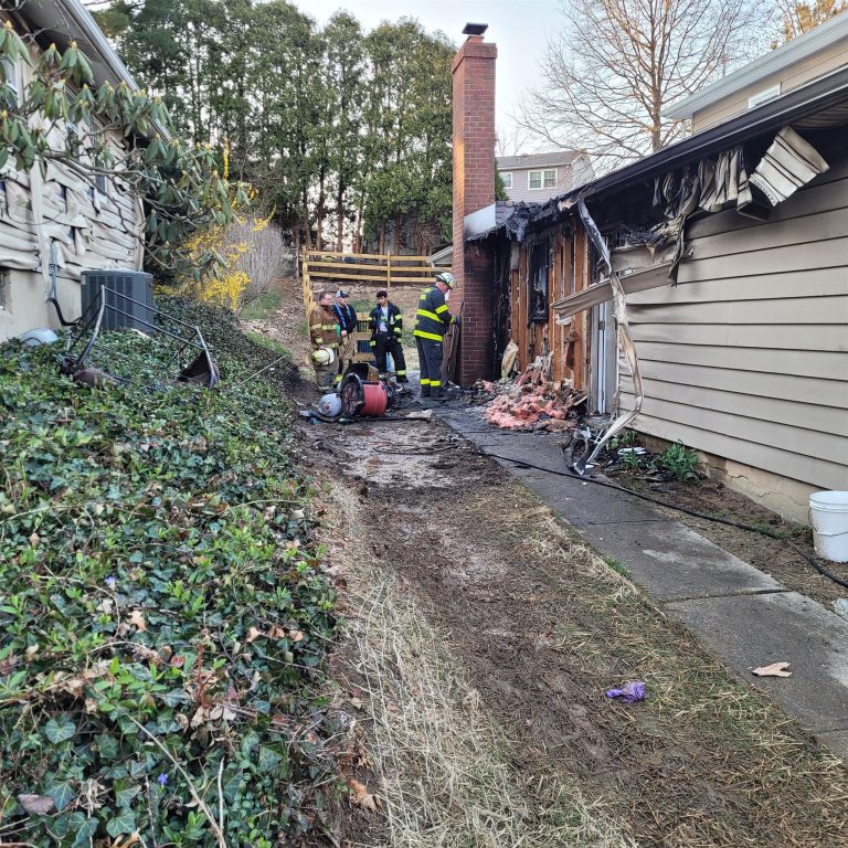 Firefighters survey the damage outside a house with visible fire damage. The siding is charred, and debris is scattered around. A few trees and bushes are in the surroundings. A large ventilation fan is on the ground nearby. The sky is clear.