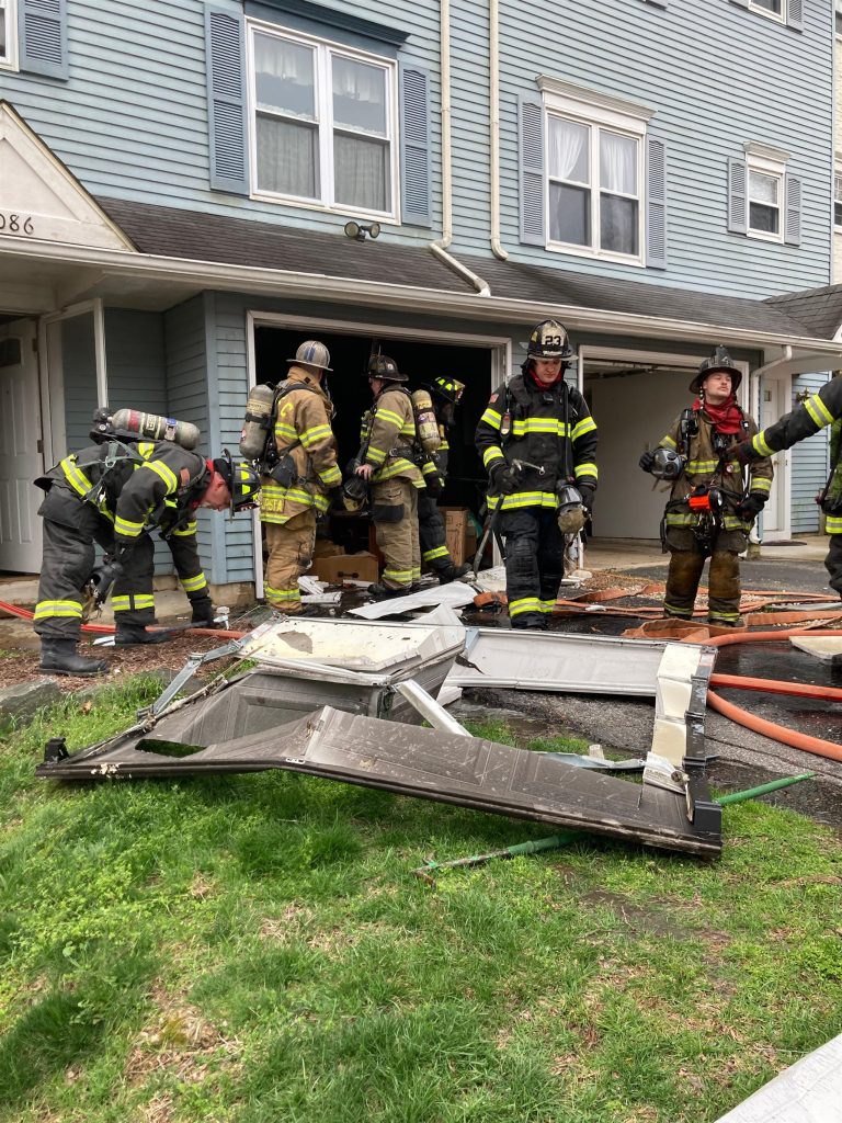 Firefighters in full gear are at the scene of a residential building incident. Casualties of broken windows and debris lie in front of the building. Some firefighters are seen working near the entrance, while others stand nearby. Hoses are scattered on the ground.