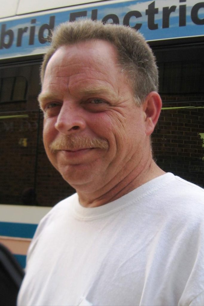 A man with short hair and a mustache, wearing a white t-shirt, is smiling slightly while standing outside. Behind him, a partially visible vehicle has the words "Hybrid Electri" on it. A brick wall and a window are in the background.