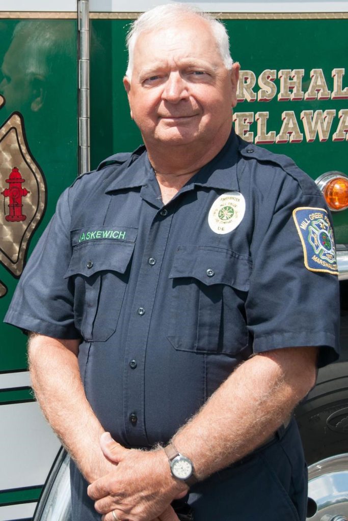 A man in a dark uniform stands in front of a green vehicle with emergency service insignias. He has short gray hair, is slightly smiling, and has his hands rested in front of him. The vehicle behind him features text mentioning "Delaware.
