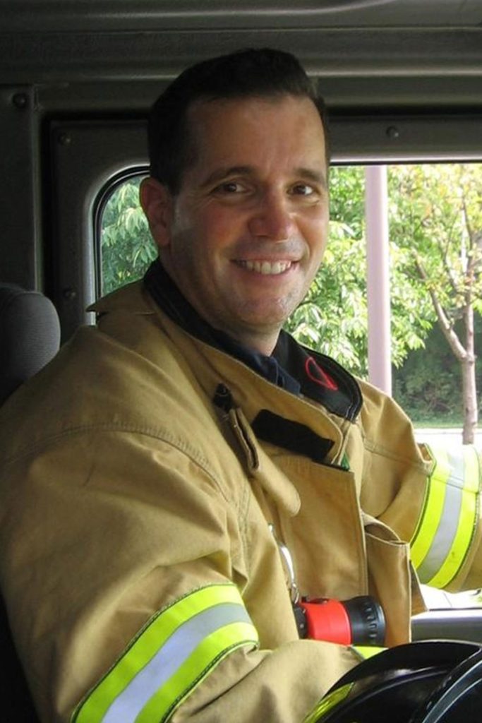 A smiling firefighter in a beige uniform with yellow reflective stripes sits in the driver's seat of a fire truck. The window in the background reveals green trees and a lamp post outside.
