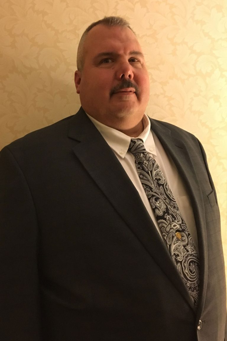 A man with short hair and a mustache is wearing a dark suit, a white shirt, and a paisley-patterned tie. He is standing in front of a beige, patterned wallpaper and looking slightly to the side.
