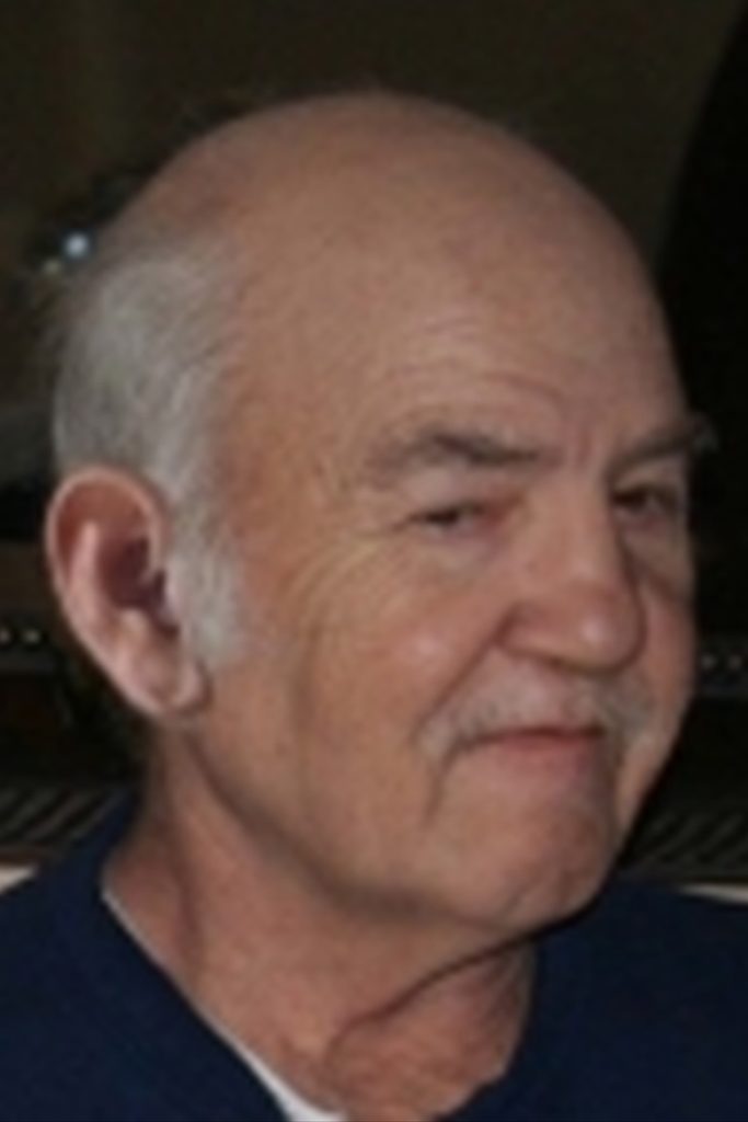 A close-up of an older man with a receding hairline and white hair. He has a slight smile and is wearing a dark shirt. The background is out of focus.
