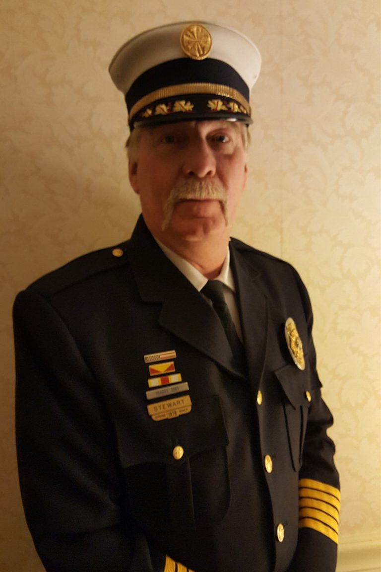 A man with a mustache wearing a formal uniform and cap with insignia and medals. The uniform features black and gold elements, and the cap has a gold emblem on the front. He stands against a patterned background in a well-lit setting.