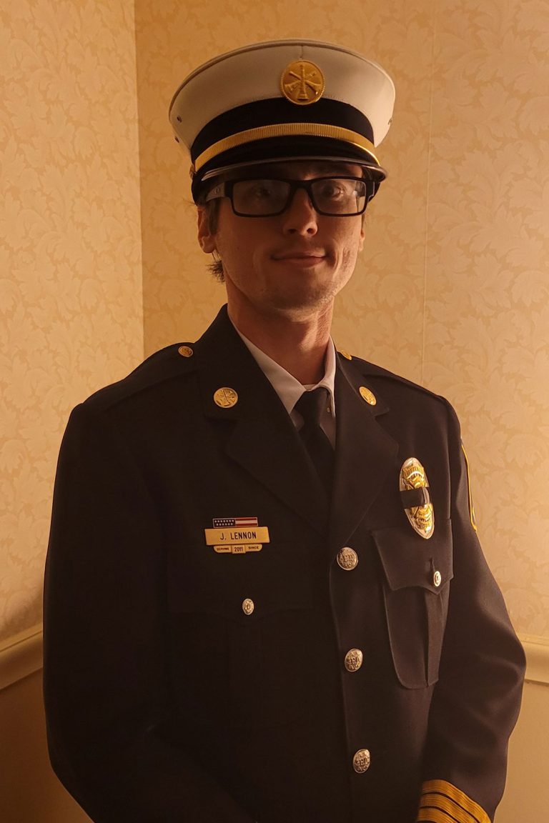 A person dressed in a formal firefighter or military uniform is standing indoors. They are wearing a white peaked cap with a gold emblem, glasses, and a jacket with insignia, including a name tag that reads "J. LENNON.