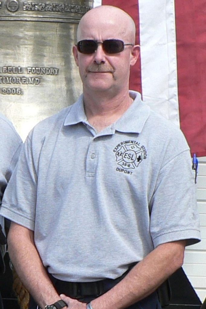 A person with a bald head, wearing dark sunglasses, and a light gray polo shirt with an embroidered emblem on the chest. The background features a large bell and part of an American flag. The individual is standing with their hands clasped in front.