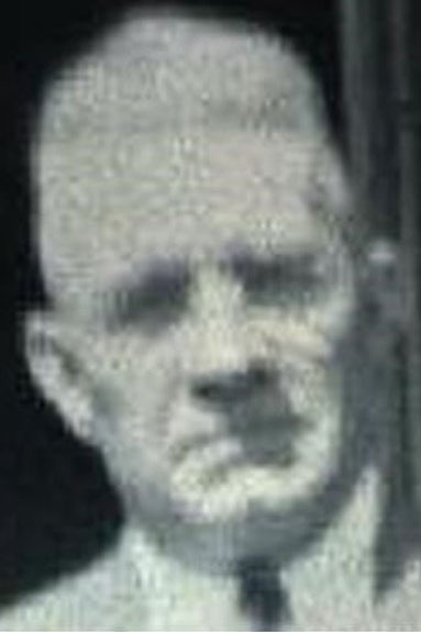 A grayscale, slightly blurred photograph of an older man with a stern expression, short-cropped hair, and wearing a suit jacket over a collared shirt. The background is dark and indistinct.