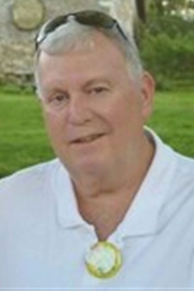 A man with short gray hair, glasses on his head, wearing a white polo shirt and a round badge or button around his neck. He is outdoors with green grass and a blurred background.