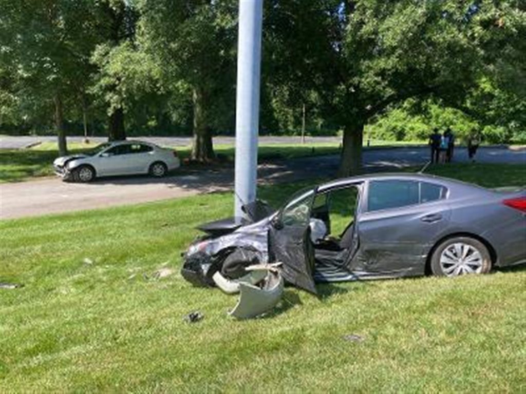 A silver sedan with significant front-end damage has collided with a light pole on a grassy area. The front bumper is detached, and the driver-side door is open. Another white car with front-end damage is parked on a nearby road. Trees and people are in the background.