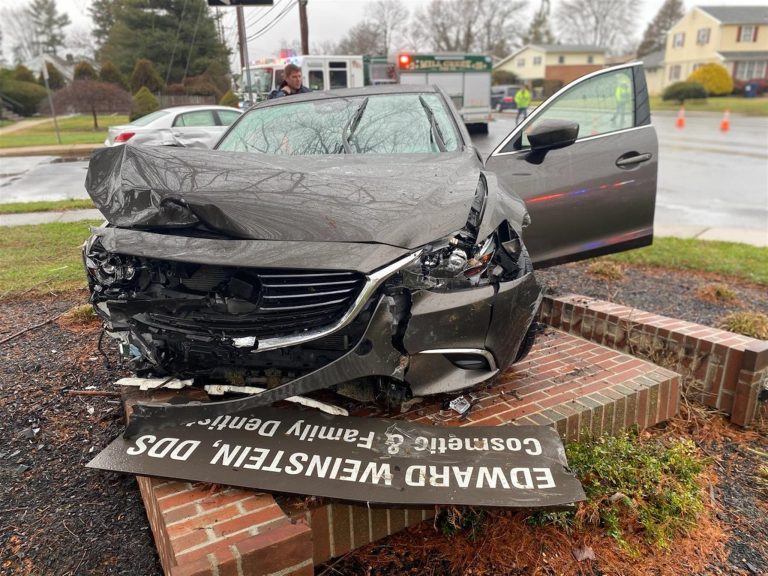 A heavily damaged car with a smashed front end is resting on a brick structure, with its front bumper partly dislodged. A damaged sign lies in front of the car. The driver's door is open, and emergency responders and a fire truck are visible in the background.