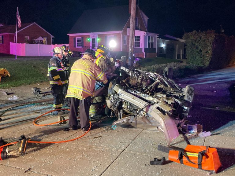 Firefighters and emergency responders work at night to rescue occupants from an overturned car on a sidewalk. The car is severely damaged, and emergency equipment such as hoses and tools are scattered around. Nearby houses are illuminated by emergency lights.