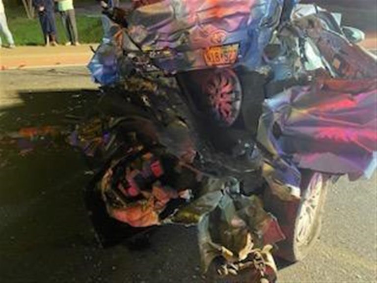 A heavily damaged vehicle with crumpled metal and debris is shown after a serious accident at night. The car’s rear end is particularly smashed, with parts scattered on the road. The rear license plate is visible. Emergency lights illuminate the scene, and people are in the background.