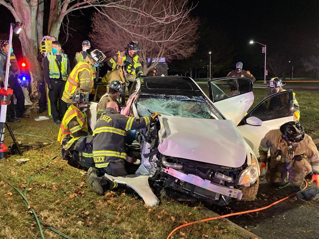 A team of firefighters in protective gear is seen at night working to extricate a person from a white car involved in a serious accident. The car's front end is heavily damaged, and the windshield is shattered. Emergency lights illuminate the scene.