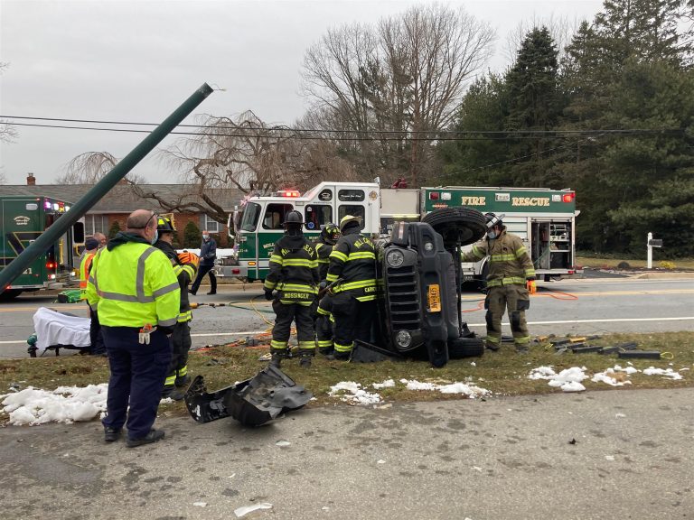 Firefighters and rescue personnel at the scene of an accident where a vehicle is flipped onto its side, next to a fallen utility pole on a roadside. Emergency vehicles and equipment are visible in the background, with snow patches on the ground.