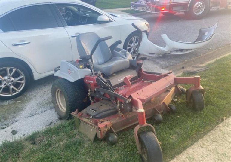 A white car with a damaged front bumper is parked at the edge of a road. In front of the car is a red riding lawn mower with some dirt on its wheels, possibly indicating it was involved in a collision. A fire truck is visible in the background.