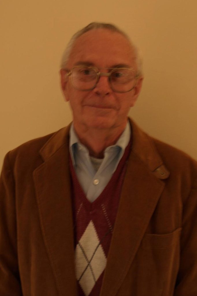 An elderly man wearing glasses, a blue collared shirt, and a maroon argyle-patterned sweater under a brown blazer stands in front of a plain beige background. He has short, gray hair and a slight smile.