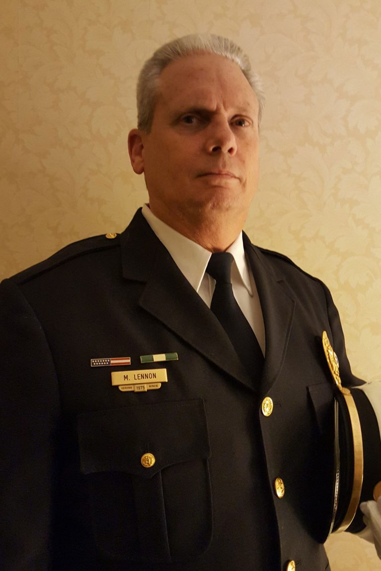 A man in a formal black military or law enforcement uniform stands against a beige patterned wall. He has short, gray hair and holds a white-capped hat under his arm. The nametag on his uniform reads "M. Lennon.