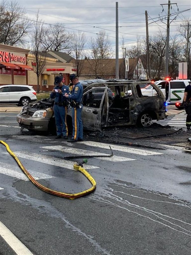 A burnt car sits at an intersection with significant fire damage. Two police officers in blue uniforms stand near the wreck, talking. A CVS Pharmacy is visible in the background. A fire hose lies on the wet road surface, and police lights flash in the distance.