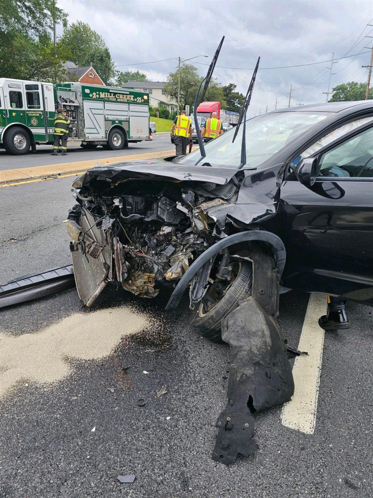 A black car with severe front-end damage is stopped on a road. Debris and parts of the car are scattered on the ground. A fire truck and a tow truck are visible in the background, as well as a firefighter and another person wearing high-visibility jackets.