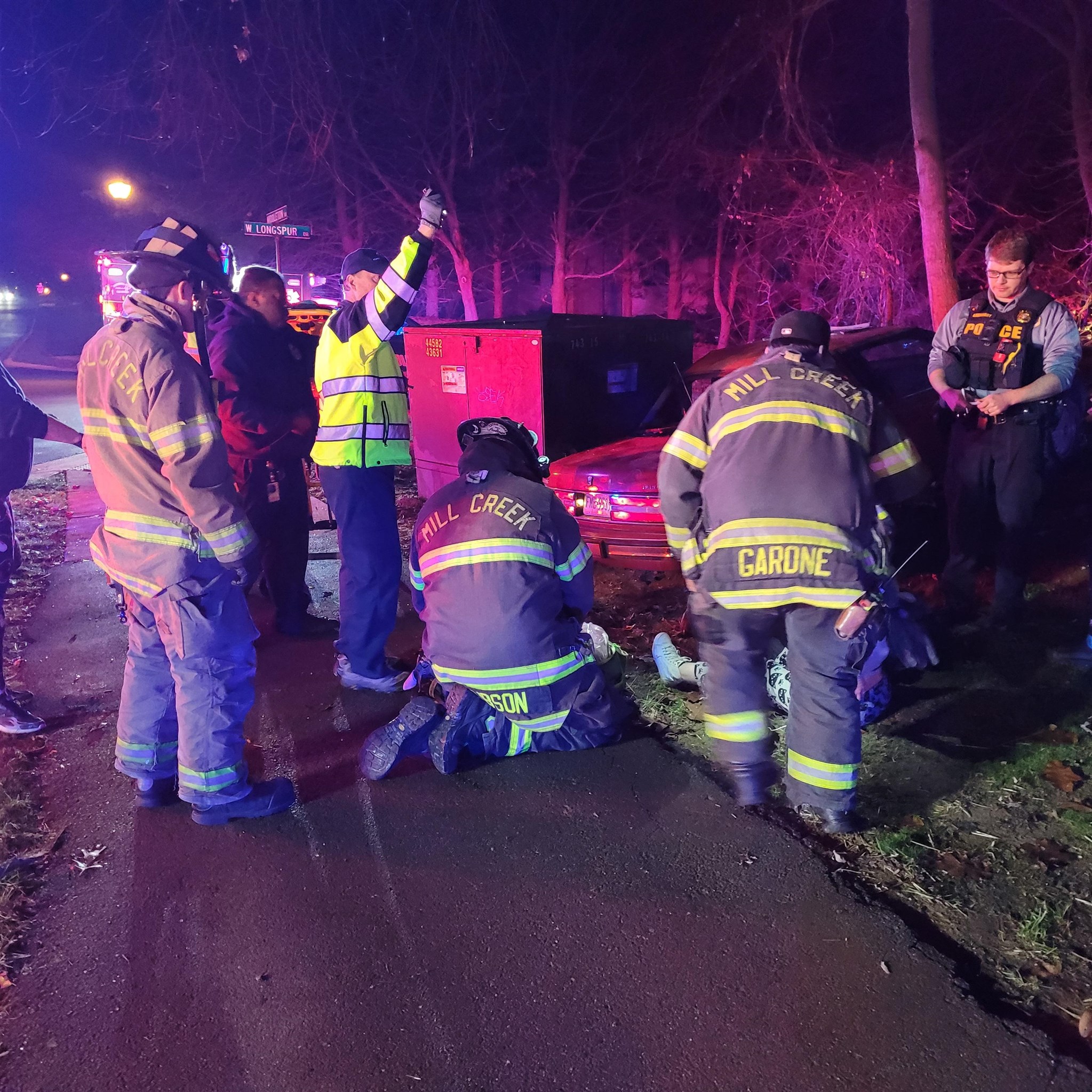 Firefighters provide assistance at the scene of a car accident at night. They are wearing reflective gear labeled with "Mill Creek." Some are kneeling beside a person on the ground, while others are standing and observing. Emergency lights illuminate the scene.