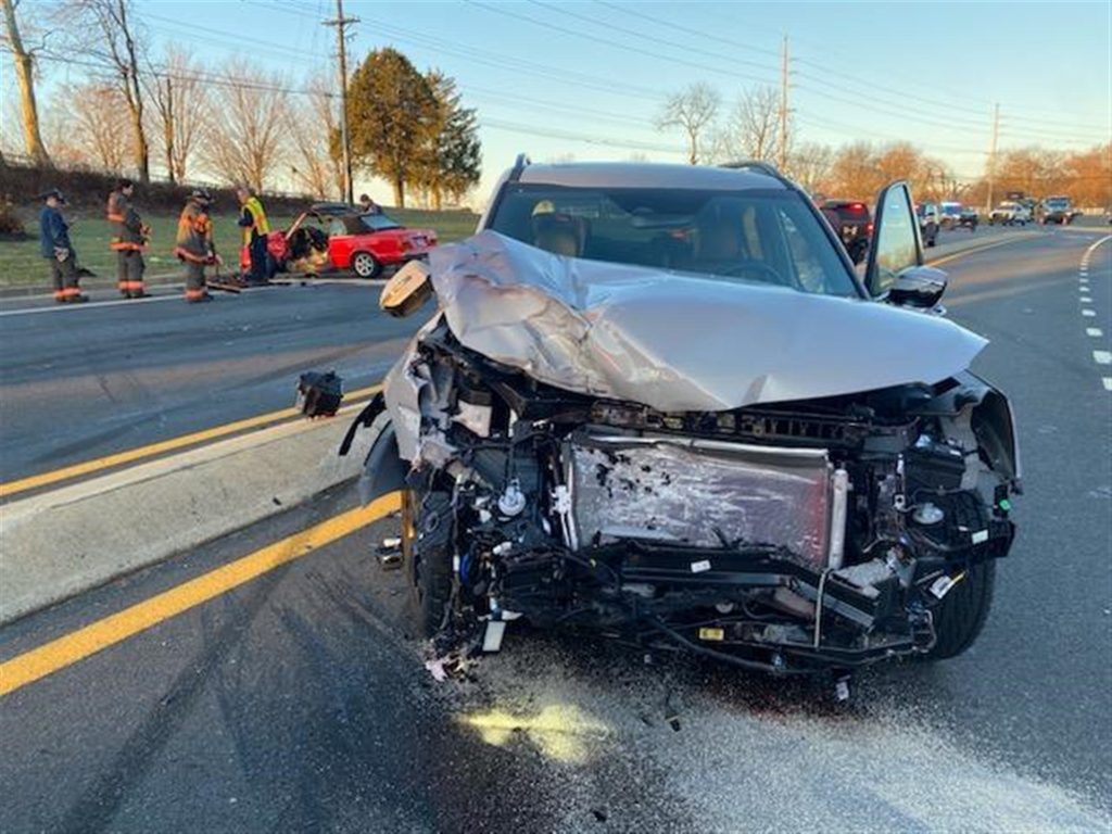 A heavily damaged SUV with a crumpled front end sits on the road next to a concrete divider. Emergency responders and bystanders are near another car, which is also damaged, in the background. Debris is scattered on the road, indicating a recent accident.