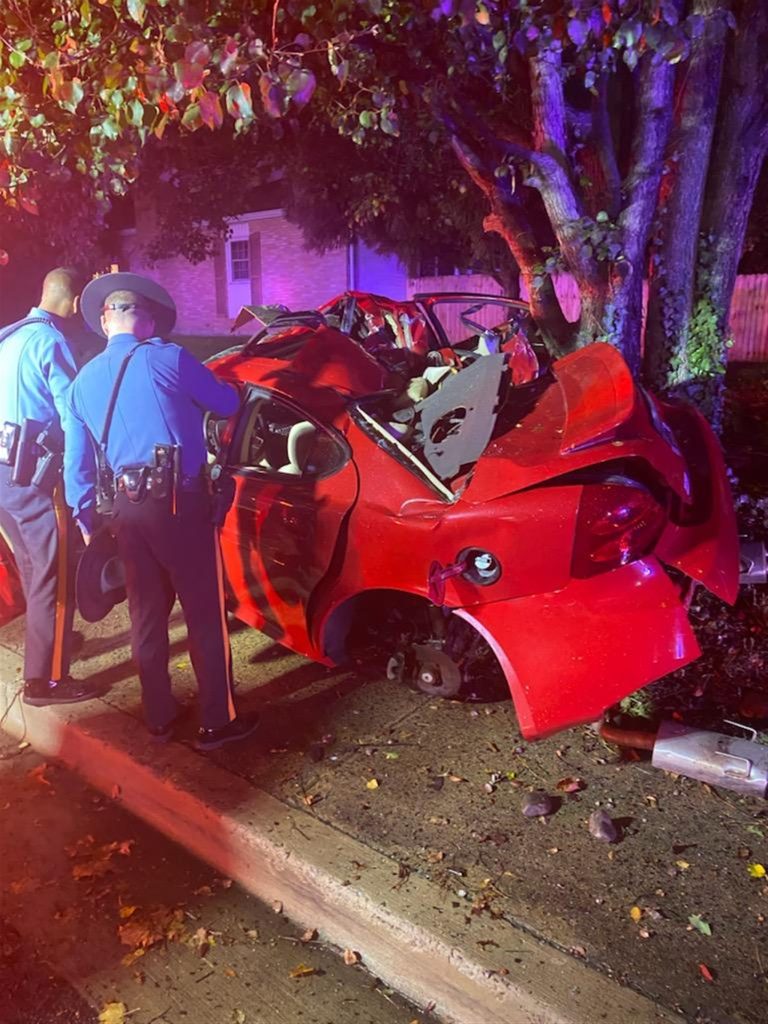 Two police officers stand next to the wreckage of a red car that appears to have crashed into a tree. The car is severely damaged, with the roof and rear end crushed. The scene is illuminated by emergency lights, and the road is wet, suggesting it may have rained recently.