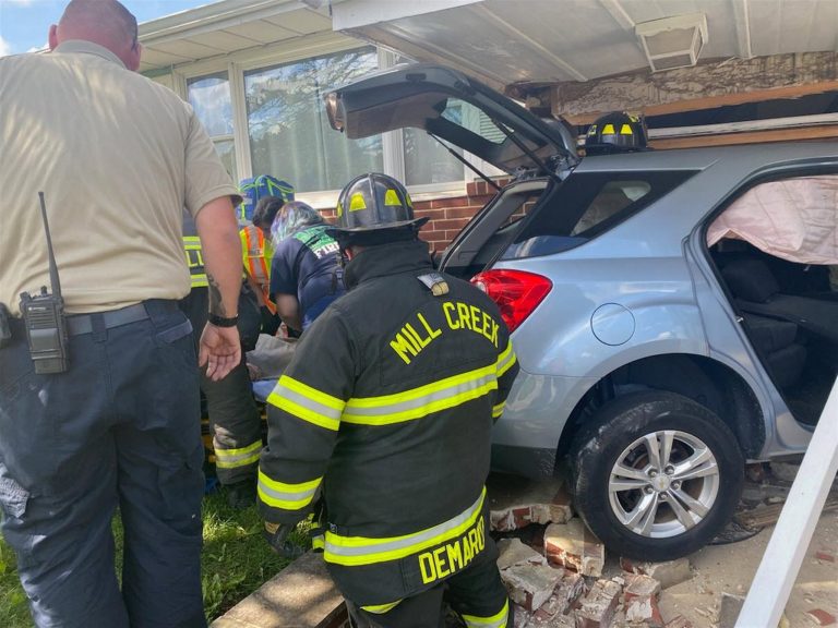 Firefighters work at the scene where an SUV has crashed into a house. One firefighter wearing gear labeled "Mill Creek" is in the foreground, while another firefighter and a person in a beige shirt assist at the side of the vehicle. Bricks and debris are scattered.