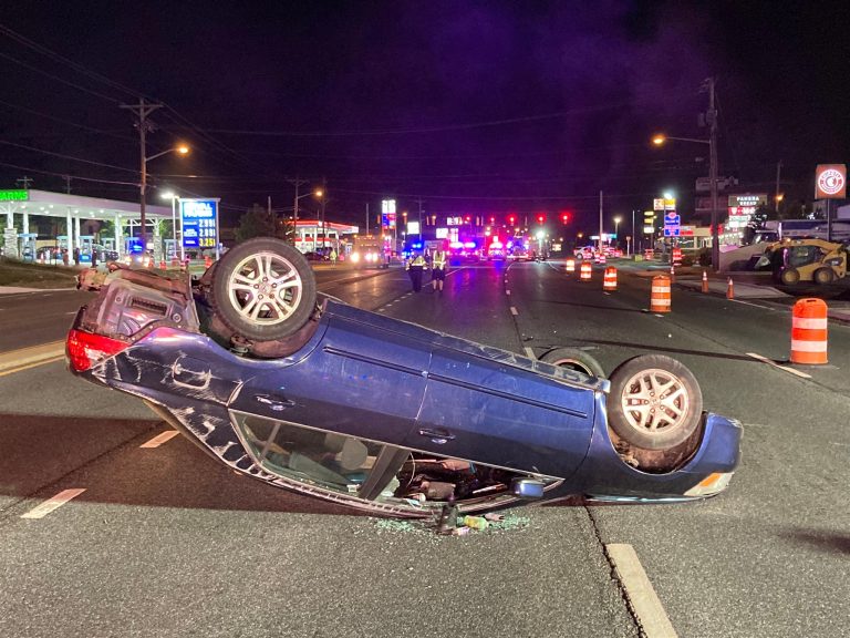 A blue car is overturned in the middle of a multi-lane road at night. Emergency vehicles with flashing lights are seen in the background, and orange traffic barrels are set up around the scene. Several storefronts with illuminated signs are visible along the road.