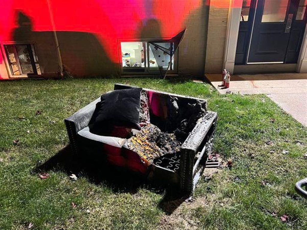 A burnt couch is placed on the grass outside a building. The couch appears heavily damaged, with charred and exposed internal materials. A fire extinguisher is seen on the ground nearby. The building's exterior wall has noticeable smoke and fire damage.