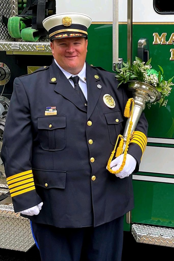 A uniformed firefighter stands proudly in front of a firetruck, wearing a decorated hat and a badge-studded jacket. He holds a traditional ceremonial trumpet adorned with a green and white floral arrangement.