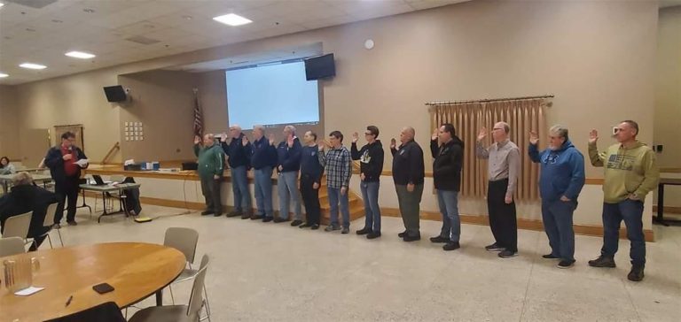 A group of people standing in a line indoors, appearing to take an oath with their right hands raised. A person to the left is addressing the group. The room contains round tables and a large projection screen on the wall behind them.