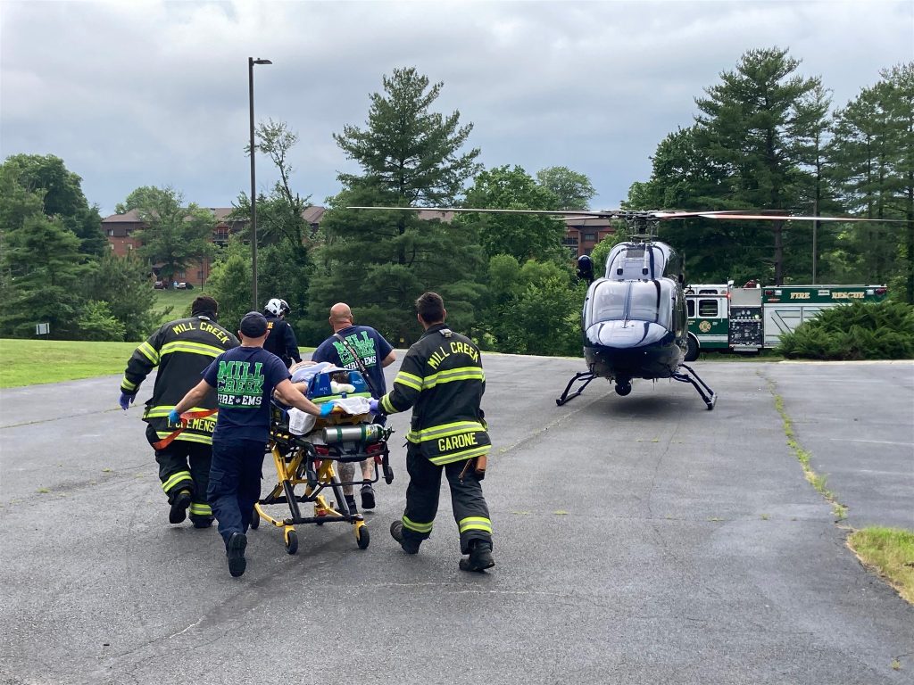 Emergency responders are transporting a person on a stretcher towards a medical helicopter parked on a road. The responders are wearing bright green and yellow reflective gear, and the scene is set outdoors with trees and emergency vehicles in the background.